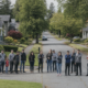 A group of neighbors and families standing in a line on a quiet suburban street, spaced apart and facing the camera.