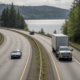 Winding highway along a lake, with a dark sedan in the left lane and a white box truck in the right lane, forested hills in the background.