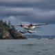 Twin-engine floatplane with pontoons landing on the sea near a rocky, forested shore; three boats nearby under cloudy skies.