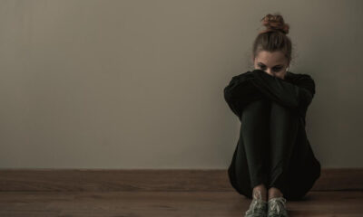 Young woman sits on a wooden floor against a beige wall, arms wrapped around her knees and looking down, conveying a somber mood.