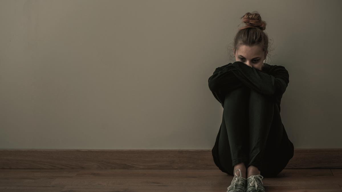 Young woman sits on a wooden floor against a beige wall, arms wrapped around her knees and looking down, conveying a somber mood.