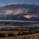 Stormy sky over jagged mountains with a bright lightning bolt striking near the peak; valley below with a small town and dark forest in foreground.