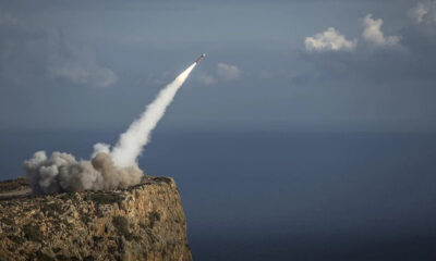 Rocket blasts off from a rocky cliff, leaving a white plume toward the sky over the ocean.