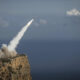 Rocket blasts off from a rocky cliff, leaving a white plume toward the sky over the ocean.