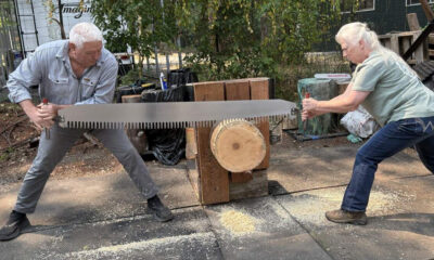Two adults share a long two-person saw to cut a wooden log placed on a wooden support outdoors, with wood chips on the ground nearby.