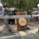 Two adults share a long two-person saw to cut a wooden log placed on a wooden support outdoors, with wood chips on the ground nearby.