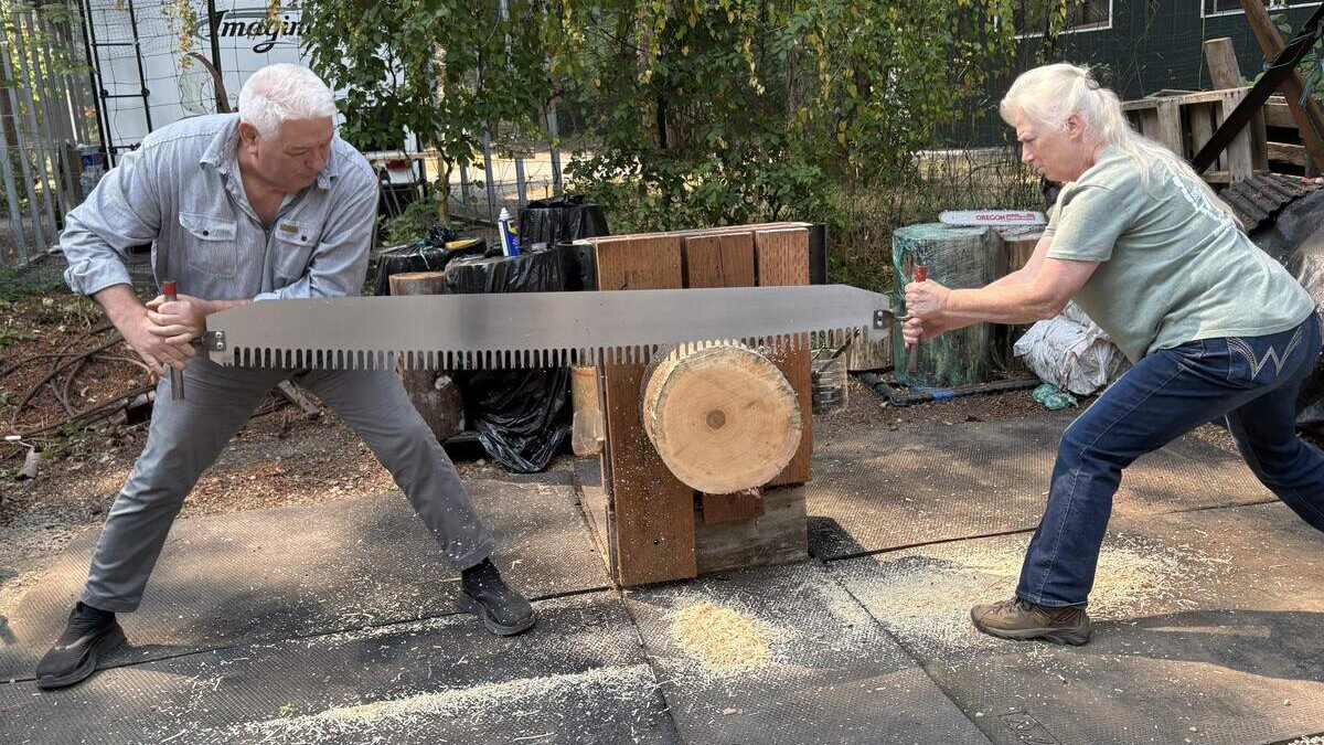 Two adults share a long two-person saw to cut a wooden log placed on a wooden support outdoors, with wood chips on the ground nearby.