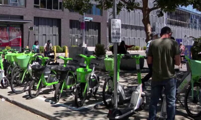Row of bright green Lime bike-share bicycles lined up outside a modern brick building, with pedestrians nearby and a scooter in view.