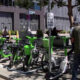 Row of bright green Lime bike-share bicycles lined up outside a modern brick building, with pedestrians nearby and a scooter in view.