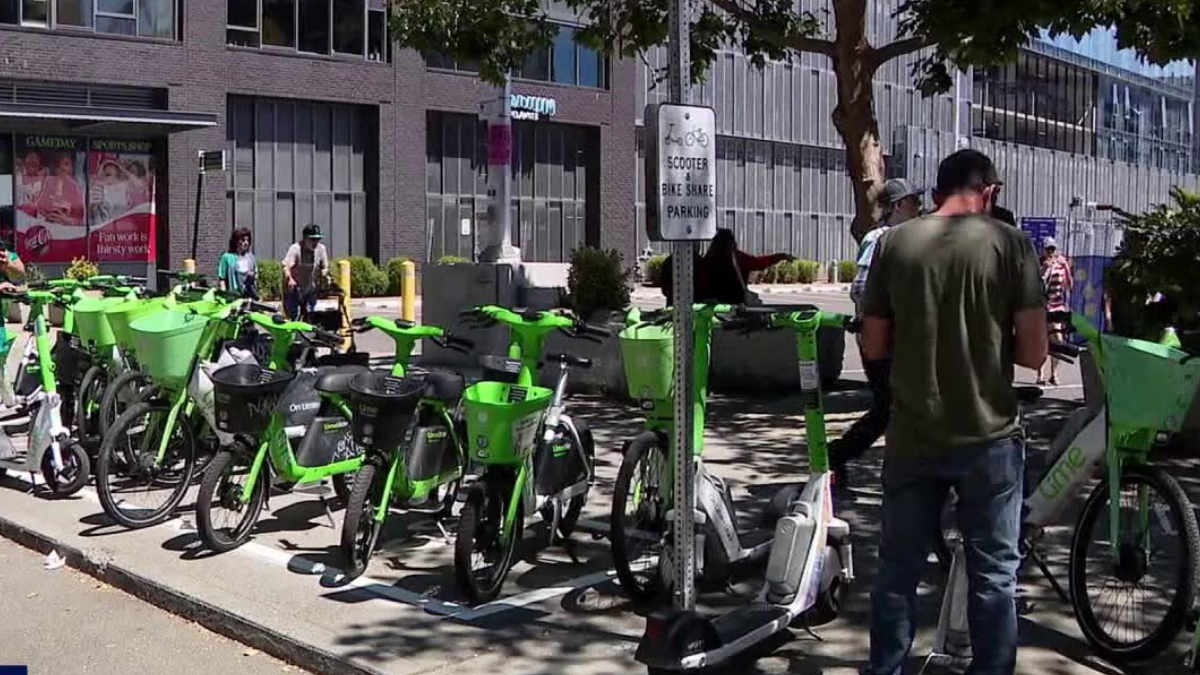 Row of bright green Lime bike-share bicycles lined up outside a modern brick building, with pedestrians nearby and a scooter in view.