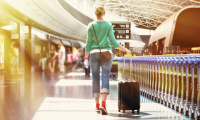 Traveler walking through an airport concourse, pulling a rolling suitcase beside a blue railing.