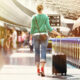 Traveler walking through an airport concourse, pulling a rolling suitcase beside a blue railing.