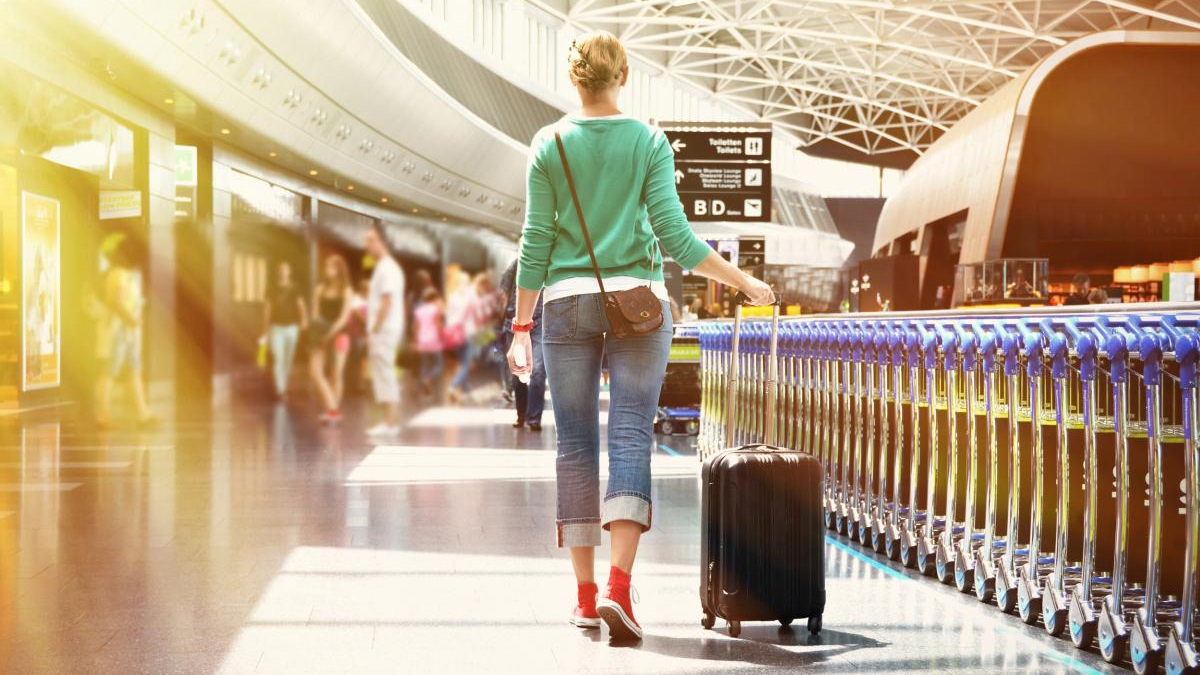 Traveler walking through an airport concourse, pulling a rolling suitcase beside a blue railing.